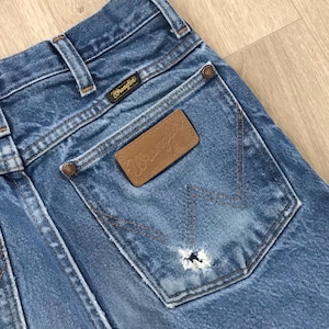 May include: Blue denim jeans featuring a brown leather patch with the Wrangler logo on the back pocket. The pocket displays a signature W-shaped stitching pattern and a small frayed hole. The jeans include a black Wrangler brand tag.