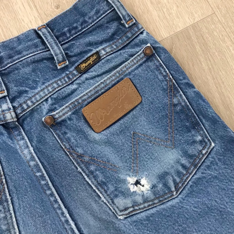 May include: Blue denim jeans featuring a brown leather patch with the Wrangler logo on the back pocket. The pocket displays a signature W-shaped stitching pattern and a small frayed hole. The jeans include a black Wrangler brand tag.