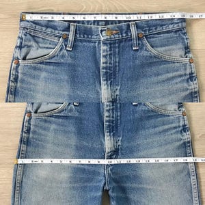 May include: Close-up of light blue denim jeans with a button and zipper closure. The jeans have front and back pockets, and copper-colored rivets. The waist measures approximately 40.6 cm across. The jeans are vintage.