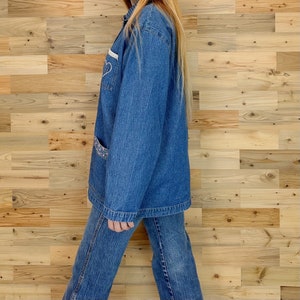 May include: A denim jacket and jeans set. The jacket features a white heart design and a pocket with a patterned fabric. The outfit is a medium wash blue color. The model is wearing the jacket and jeans.