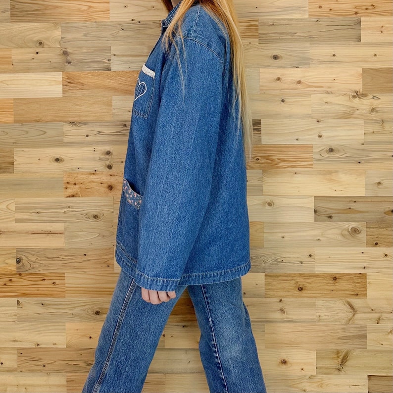 May include: A denim jacket and jeans set. The jacket features a white heart design and a pocket with a patterned fabric. The outfit is a medium wash blue color. The model is wearing the jacket and jeans.
