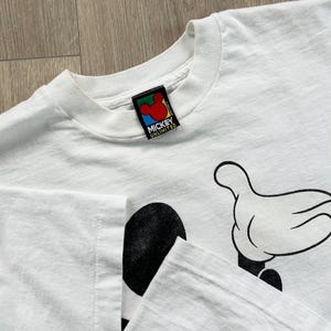 Op de afbeelding: Wit T-shirt met een Mickey Mouse-afbeelding, met zijn hand en een deel van zijn gezicht. Het shirt heeft een ronde hals en een label met de tekst "Mickey Unlimited". De afbeelding is zwart en wit.