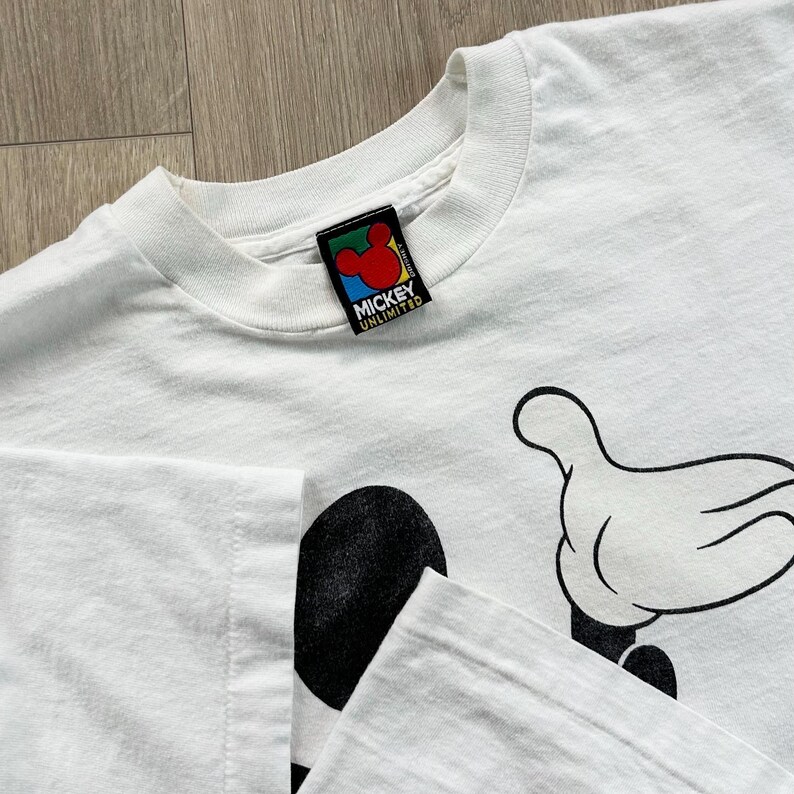 Op de afbeelding: Wit T-shirt met een Mickey Mouse-afbeelding, met zijn hand en een deel van zijn gezicht. Het shirt heeft een ronde hals en een label met de tekst "Mickey Unlimited". De afbeelding is zwart en wit.