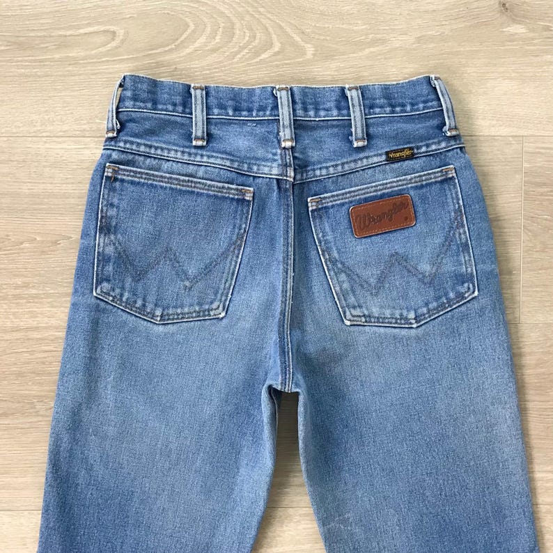 Op de afbeelding: Lichtblauwe spijkerbroek met een klassiek ontwerp met vijf zakken. De jeans heeft een bruine leren patch met het "Wrangler"-logo op de achterzak. De achterzakken hebben een kenmerkende "W"-stitching.