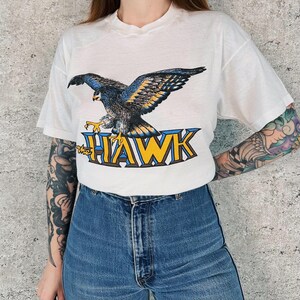 May include: White t-shirt featuring a detailed graphic of a hawk with blue, yellow, and black feathers. The text "the HAWK" is printed below the hawk in blue and yellow. The shirt is paired with blue jeans.