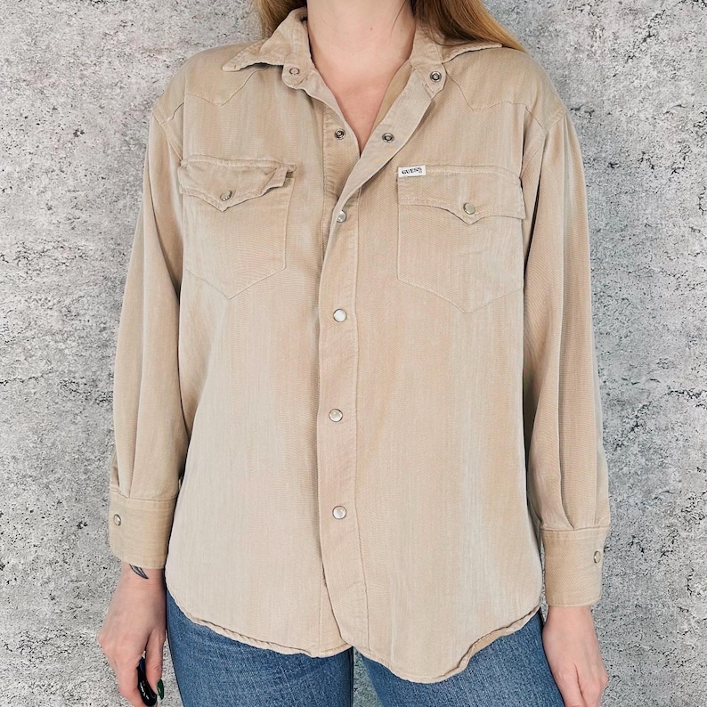 May include: A tan button-down shirt with long sleeves, two chest pockets, and a collar. The shirt has a small "Vans" label and is made of a corduroy-like material. The shirt is worn with blue jeans.