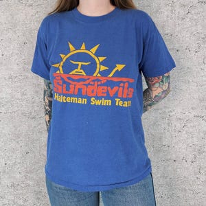 May include: Blue t-shirt featuring a sun graphic with the text "Sundevils Halteman Swim Team" in orange and white. The shirt has short sleeves and a crew neck.
