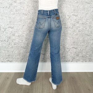 May include: Light blue denim jeans with a classic fit. The jeans feature a high waist, back pockets with a 'W' stitch design, and a brown leather patch. The jeans have a slight flare at the hem.