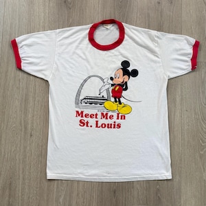May include: White t-shirt with red trim featuring a graphic of Mickey Mouse in front of a St. Louis landmark. The text "Meet Me In St. Louis" is printed in red below the graphic. The shirt has short sleeves.