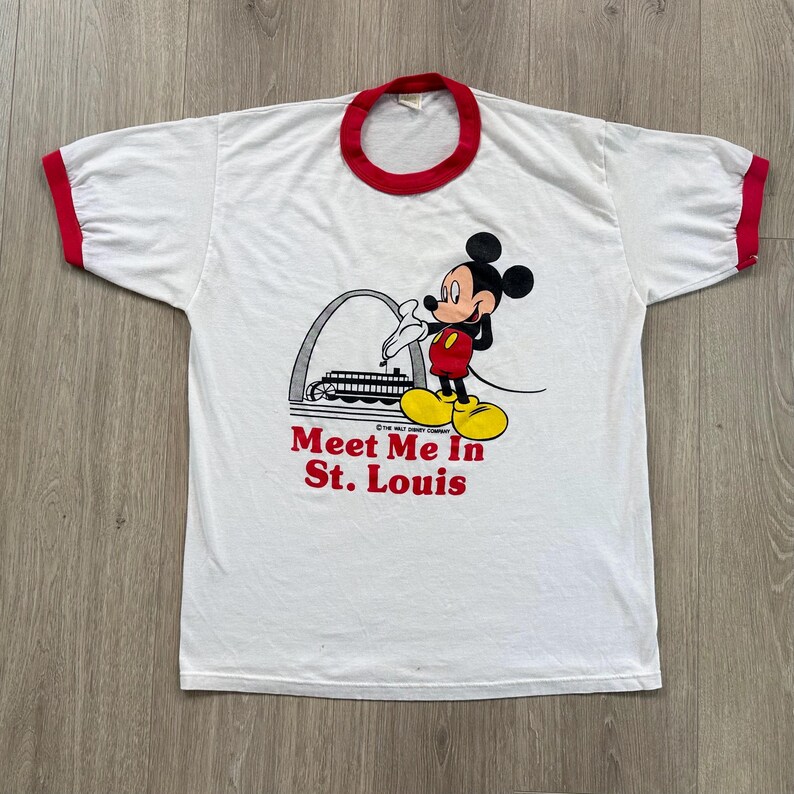 May include: White t-shirt with red trim featuring a graphic of Mickey Mouse in front of a St. Louis landmark. The text "Meet Me In St. Louis" is printed in red below the graphic. The shirt has short sleeves.