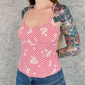 May include: A pink and white gingham print tank top with white daisy floral embroidery. The top has thin straps and a scoop neckline. The model is wearing blue jeans.