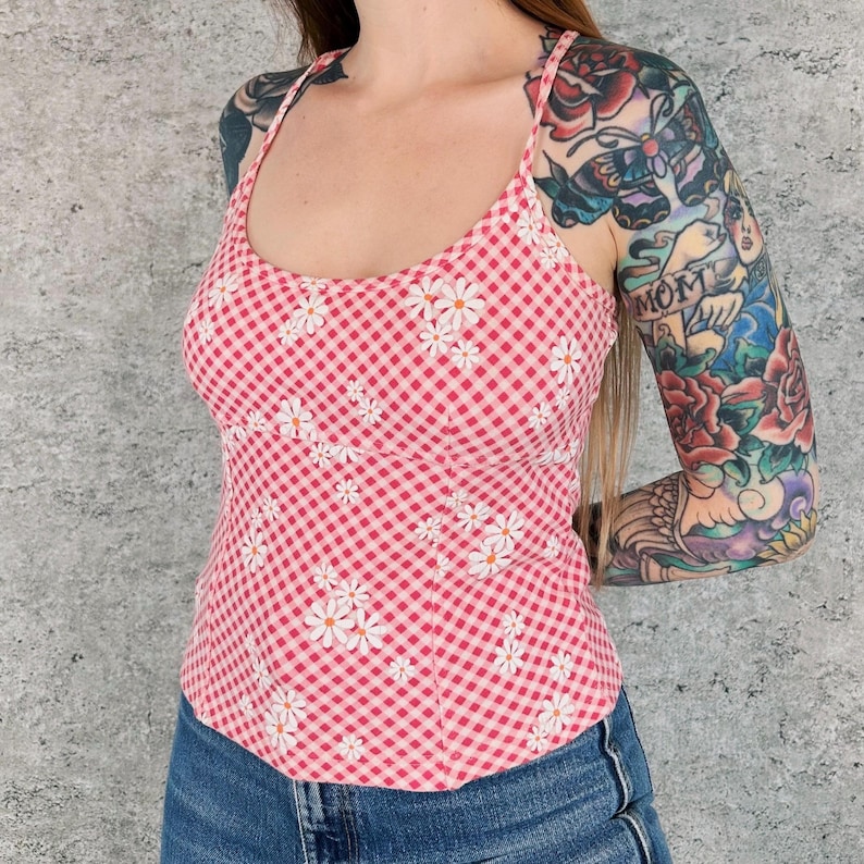 May include: A pink and white gingham print tank top with white daisy floral embroidery. The top has thin straps and a scoop neckline. The model is wearing blue jeans.
