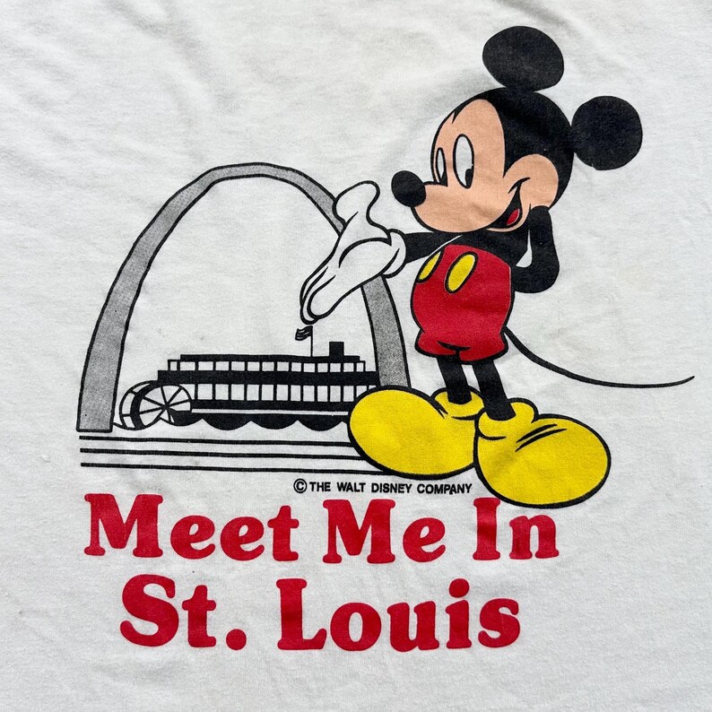 May include: White t-shirt featuring Mickey Mouse with the Gateway Arch and a steamboat. The text "Meet Me In St. Louis" is printed in red. The Walt Disney Company logo is also visible.