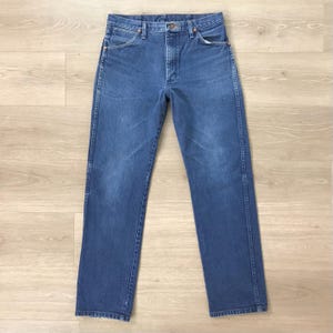 K&ouml;nnte beinhalten: Eine blaue Jeanshose mit klassischem F&uuml;nf-Taschen-Design. Die Jeans hat einen geraden Beinverlauf und ist aus mittelblauem Denim gefertigt. Die Jeans wird auf einer hellen Holzoberfl&auml;che pr&auml;sentiert.