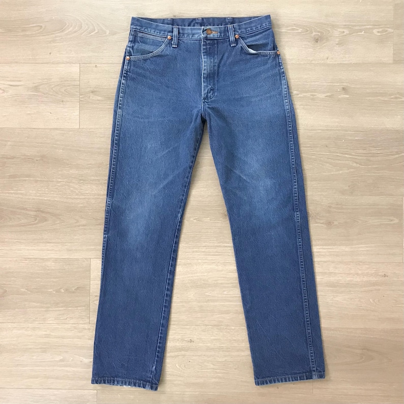K&ouml;nnte beinhalten: Eine blaue Jeanshose mit klassischem F&uuml;nf-Taschen-Design. Die Jeans hat einen geraden Beinverlauf und ist aus mittelblauem Denim gefertigt. Die Jeans wird auf einer hellen Holzoberfl&auml;che pr&auml;sentiert.