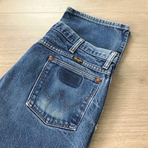 Puede incluir: Primer plano de unos vaqueros de mezclilla azules. Los vaqueros presentan un dise&ntilde;o cl&aacute;sico de cinco bolsillos, con una etiqueta Wrangler en el bolsillo trasero. Los vaqueros est&aacute;n doblados, mostrando los bolsillos traseros y la cintura. La mezclilla tiene un aspecto descolorido y desgastado.