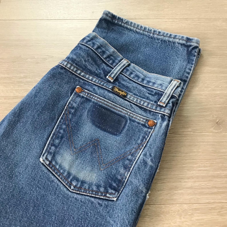 Puede incluir: Primer plano de unos vaqueros de mezclilla azules. Los vaqueros presentan un dise&ntilde;o cl&aacute;sico de cinco bolsillos, con una etiqueta Wrangler en el bolsillo trasero. Los vaqueros est&aacute;n doblados, mostrando los bolsillos traseros y la cintura. La mezclilla tiene un aspecto descolorido y desgastado.