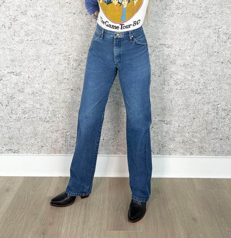K&ouml;nnte beinhalten: Blaue Jeans aus Denim mit klassischem geradem Bein. Die Jeans haben eine mittelblaue Waschung und ein traditionelles F&uuml;nf-Taschen-Design. Das Model tr&auml;gt schwarze Lederstiefel. Das Oberteil hat den Text "The Game Tour 80".