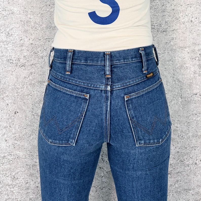 May include: Close-up of the back of blue denim jeans with the Wrangler logo on the back pocket. The jeans have a classic five-pocket design and a high waist. The person is wearing a cream-colored shirt with a blue design.