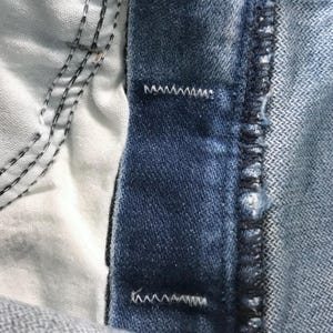 May include: A close-up view of blue denim fabric featuring a thick waistband with visible white zigzag stitching at the top and bottom edges. The interior of the garment shows a white pocket lining with dark blue stitching along the seams.