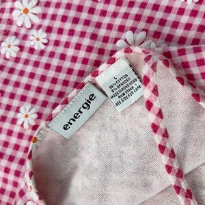 May include: Close-up of a pink and white gingham fabric with white daisy accents. The tag reads "energie" and includes fabric content and care instructions. The fabric is a blend of cotton and spandex.