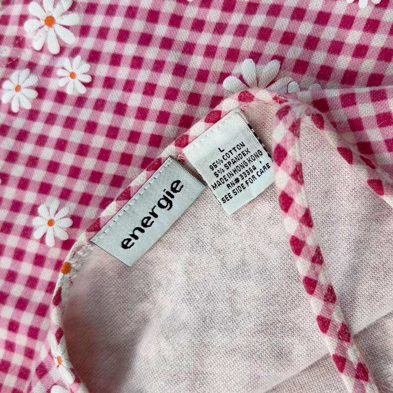 May include: Close-up of a pink and white gingham fabric with white daisy accents. The tag reads "energie" and includes fabric content and care instructions. The fabric is a blend of cotton and spandex.