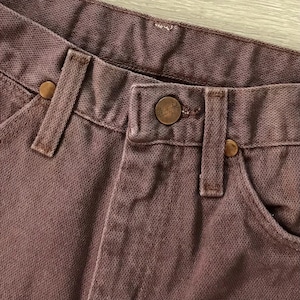 Op de afbeelding: Close-up van een bruine spijkerbroek. De jeans heeft een knoopsluiting, riemlussen en koperkleurige klinknagels. De merknaam Wrangler is zichtbaar op de knoop. De stof heeft een getextureerde uitstraling.