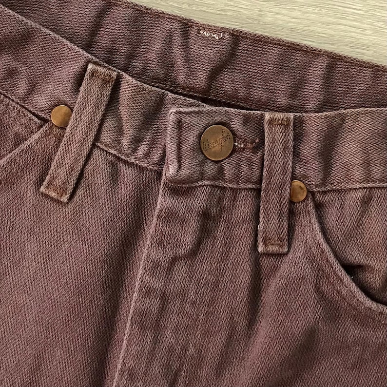 Op de afbeelding: Close-up van een bruine spijkerbroek. De jeans heeft een knoopsluiting, riemlussen en koperkleurige klinknagels. De merknaam Wrangler is zichtbaar op de knoop. De stof heeft een getextureerde uitstraling.