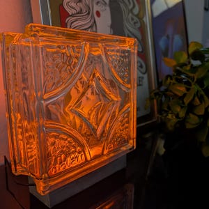 May include: A square glass block lamp emitting a warm orange glow. The front surface features a raised, textured geometric pattern with a central diamond shape surrounded by embossed floral-like details, all encased in a clear, thick glass frame.