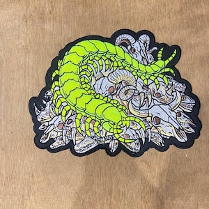 May include: An embroidered patch featuring a bright neon green centipede coiled over a cluster of grey and white animal skulls. The design is outlined in black thread, creating a high-contrast graphic illustration of a multi-legged insect.