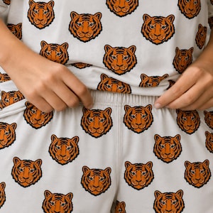 May include: A two-piece pajama set featuring a repeating pattern of orange and black tiger faces on a light cream background. The set consists of a short-sleeved top and matching shorts with an elastic waistband, showcasing a consistent tiger print.