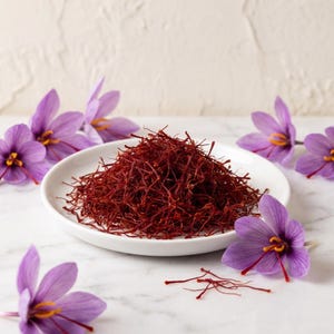 Könnte beinhalten: Ein kleiner weißer Teller, der mit dunkelroten Safranfäden angehäuft ist, umgeben von mehreren violetten Krokusblüten mit leuchtend orangefarbenen Staubgefäßen. Die feinen, dünnen Fäden des Gewürzes sind in der Mitte des glatten, runden Tellers.
