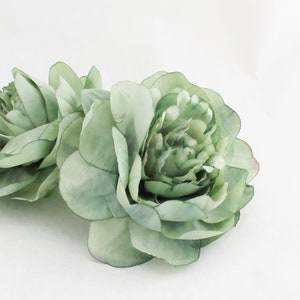 May include: Two large, light green artificial peony flowers with multiple layers of petals. The flowers have a soft, watercolor-like appearance and are set against a white background. The petals have subtle variations in color, adding depth and realism.