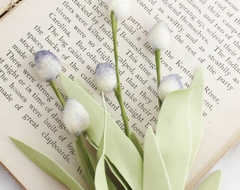 May include: A close-up of a faux floral arrangement featuring white, fuzzy buds with a hint of lavender, green stems, and light green leaves. The arrangement is set against the backdrop of an open book.