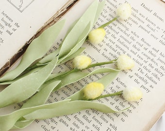 May include: A close-up of artificial yellow and white flowers with green leaves, resting on the pages of an open vintage book. The flowers have a fuzzy texture and are arranged diagonally across the page.