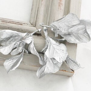 May include: Silver-painted leaf and stem arrangement, possibly for decorative purposes. The leaves have a textured appearance and are placed on a distressed, light gray wooden frame. The leaves are detailed and have a slight sparkle.