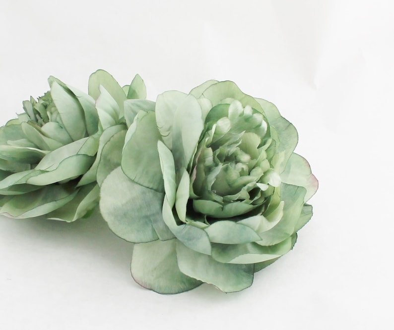 May include: Two large, light green artificial peony flowers with multiple layers of petals. The flowers have a soft, watercolor-like appearance and are set against a white background. The petals have subtle variations in color, adding depth and realism.