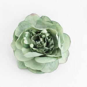 May include: A large, sage green artificial flower with multiple layers of petals. The flower has a full, rounded shape and a slightly darker center. The petals have a delicate, translucent quality and a hint of pink at the edges. The flower is set against a white background.