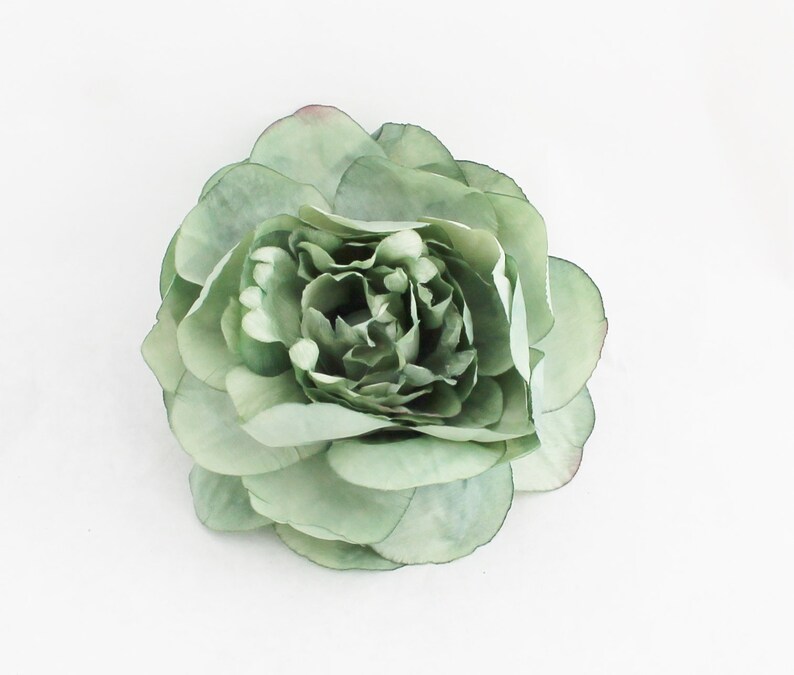 May include: A large, sage green artificial flower with multiple layers of petals. The flower has a full, rounded shape and a slightly darker center. The petals have a delicate, translucent quality and a hint of pink at the edges. The flower is set against a white background.