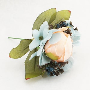 May include: A close-up of a small, artificial floral arrangement. The arrangement features a peach-colored rose surrounded by light blue flowers and green leaves. Small blue berries and dark brown accents add detail. The stem is green.