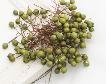 May include: A bundle of artificial berries with small, round, olive-green fruit. The berries are attached to thin, brown stems, creating a naturalistic appearance. The arrangement is set against a white background, suggesting a decorative or craft item.