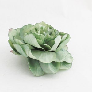 May include: A large, artificial flower in shades of green, with a hint of pink at the edges of some petals. The flower has multiple layers of petals, creating a full, textured appearance. The petals are slightly ruffled, adding to the realistic look of the floral design.