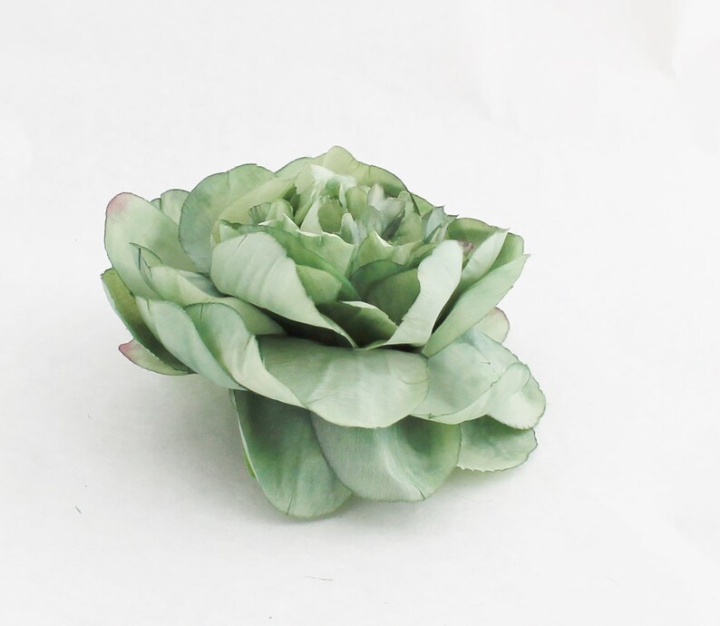 May include: A large, artificial flower in shades of green, with a hint of pink at the edges of some petals. The flower has multiple layers of petals, creating a full, textured appearance. The petals are slightly ruffled, adding to the realistic look of the floral design.
