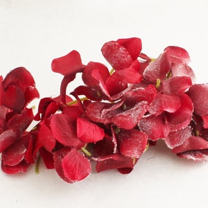May include: A cluster of deep red artificial flowers with a dusting of white powder, possibly for decoration. The petals are large and textured, with a slight sheen. The stems are green and the background is white.