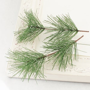 May include: Three sprigs of artificial pine branches with a frosted finish. The branches are green and have brown stems.