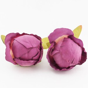 May include: Two artificial peony flower buds in a deep magenta hue. The petals are tightly furled, with a hint of a lighter color at the center. Each bud has a small, light green leaf attached, set against a white background.