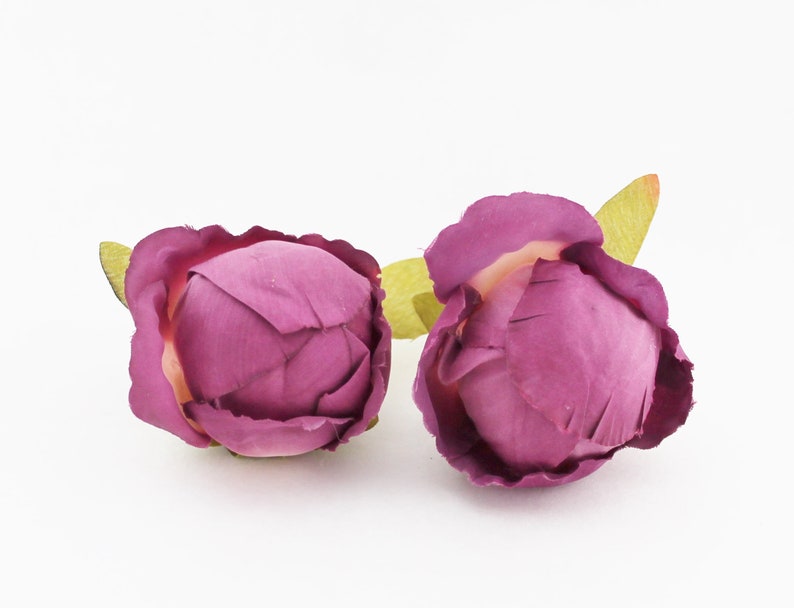 May include: Two artificial peony flower buds in a deep magenta hue. The petals are tightly furled, with a hint of a lighter color at the center. Each bud has a small, light green leaf attached, set against a white background.