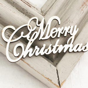 May include: White wooden cutout of the phrase "Merry Christmas" with a cursive font. The cutout is on a light gray wooden surface.