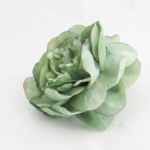 May include: A large, artificial flower with multiple layers of soft green petals. The flower has a realistic appearance, with subtle variations in color and texture. The edges of some petals have a hint of pink. The flower is likely a peony or a similar type of bloom.