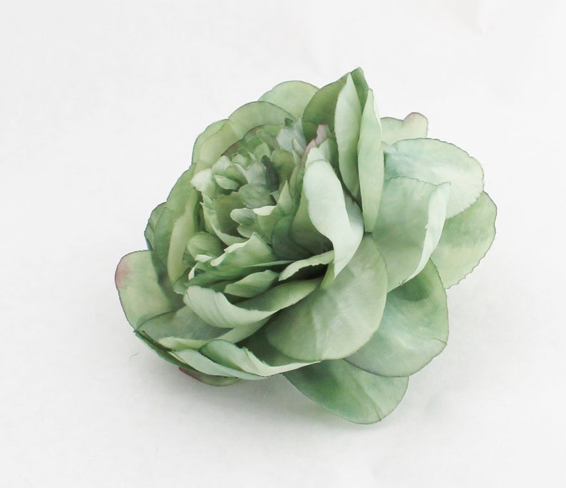 May include: A large, artificial flower with multiple layers of soft green petals. The flower has a realistic appearance, with subtle variations in color and texture. The edges of some petals have a hint of pink. The flower is likely a peony or a similar type of bloom.